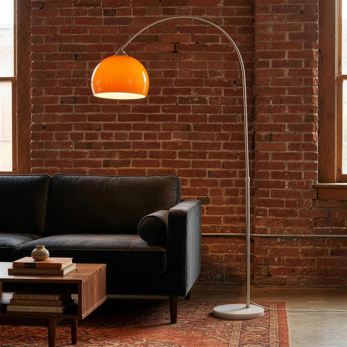 Ardo orange arc floor lamp arching over a charcoal sofa in a loft living room with exposed brick
