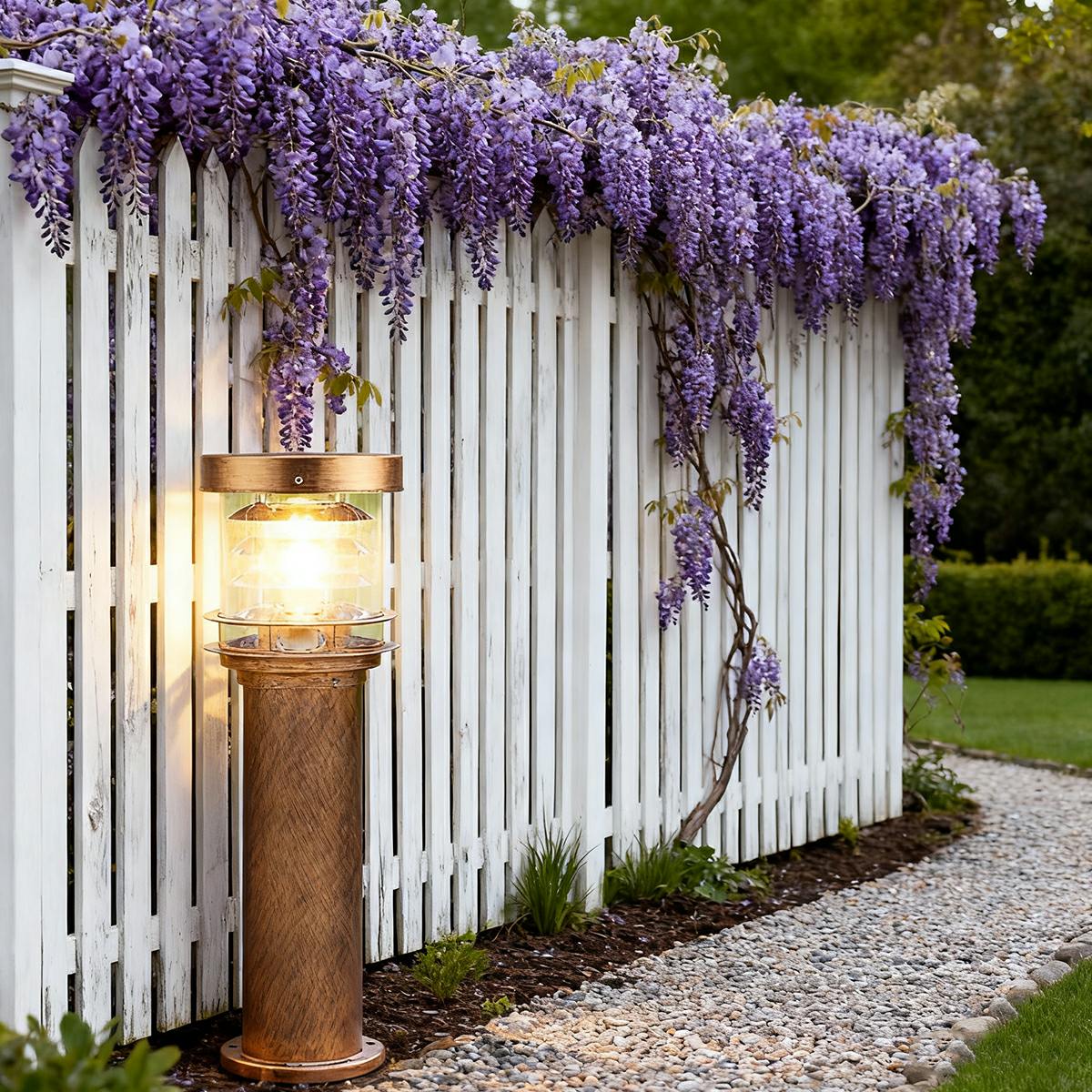 bulba bronze outdoor pillar light shining warmly next to a white garden fence and purple wisteria