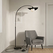 Clent black arc floor lamp with a hat-shaped shade arching over a grey armchair in a pale living room corner