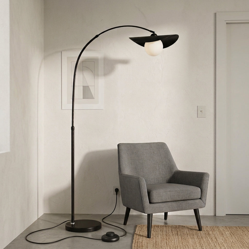 Clent black arc floor lamp with a hat-shaped shade arching over a grey armchair in a pale living room corner