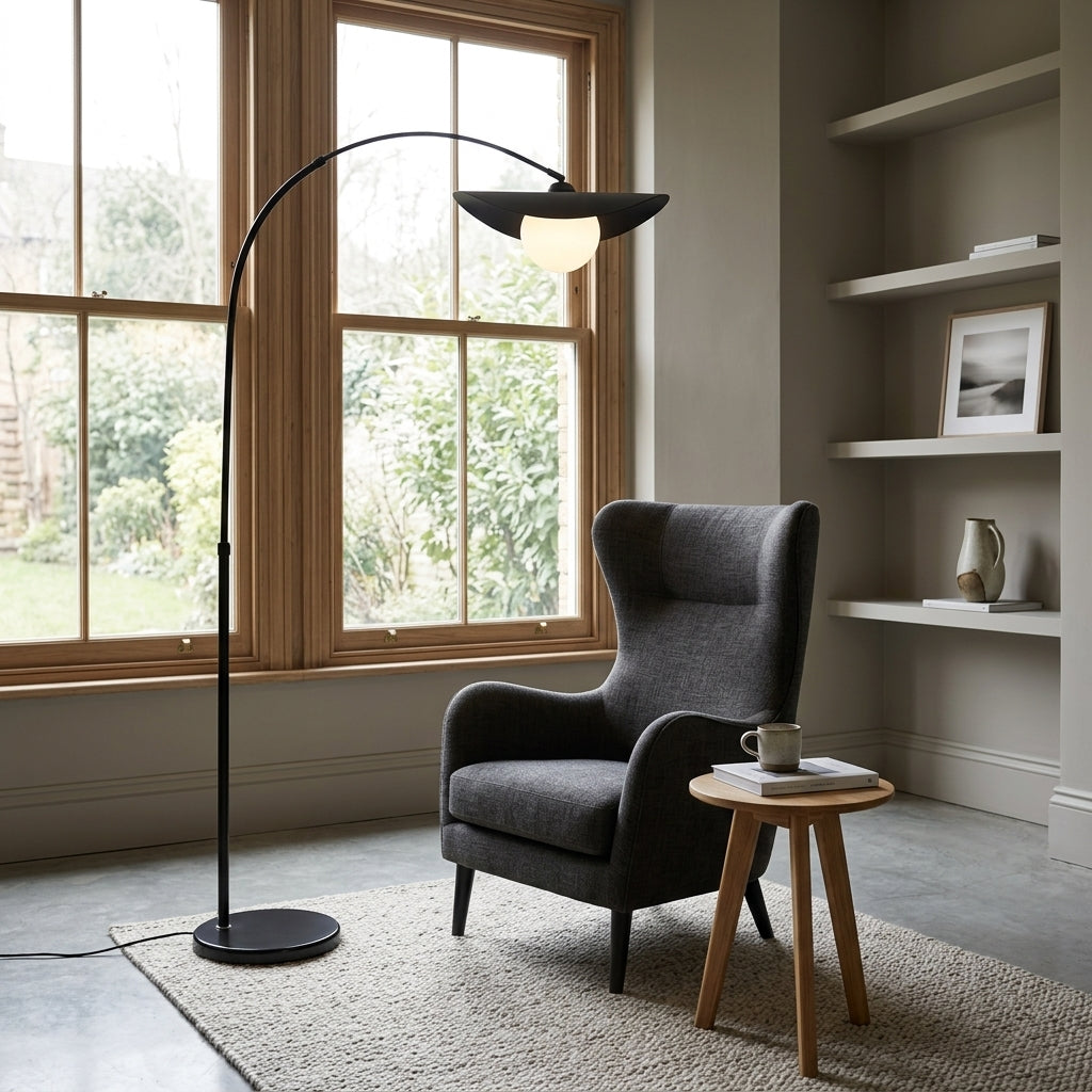 Clent black arc floor lamp beside a dark grey wing chair and a round wooden side table by a sash window