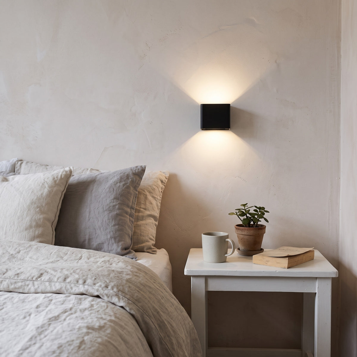 Cubo black cube wall light on a plaster bedroom wall above a white bedside table with a mug, plant and book