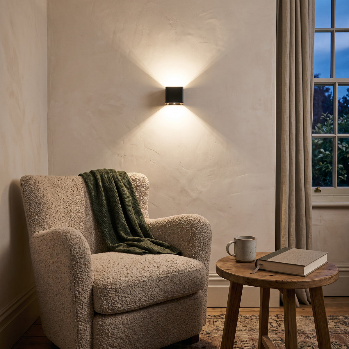 Cubo black cube wall light above a boucle armchair and wooden side table with a mug and book in a reading nook