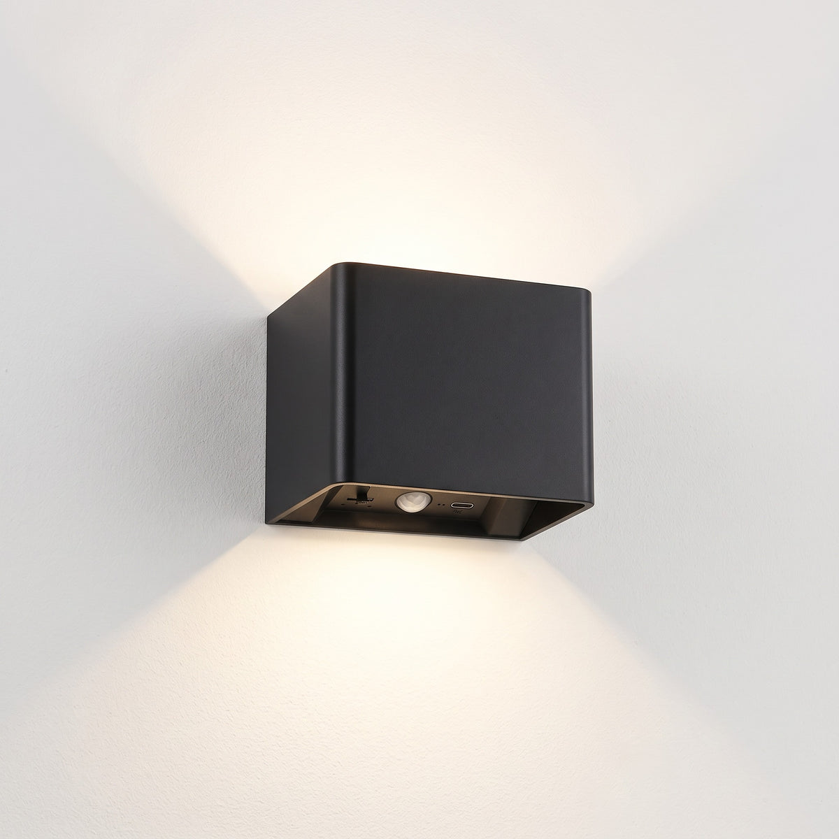 Cubo black cube wall light switched on, casting a soft up and down glow on a plain off-white wall