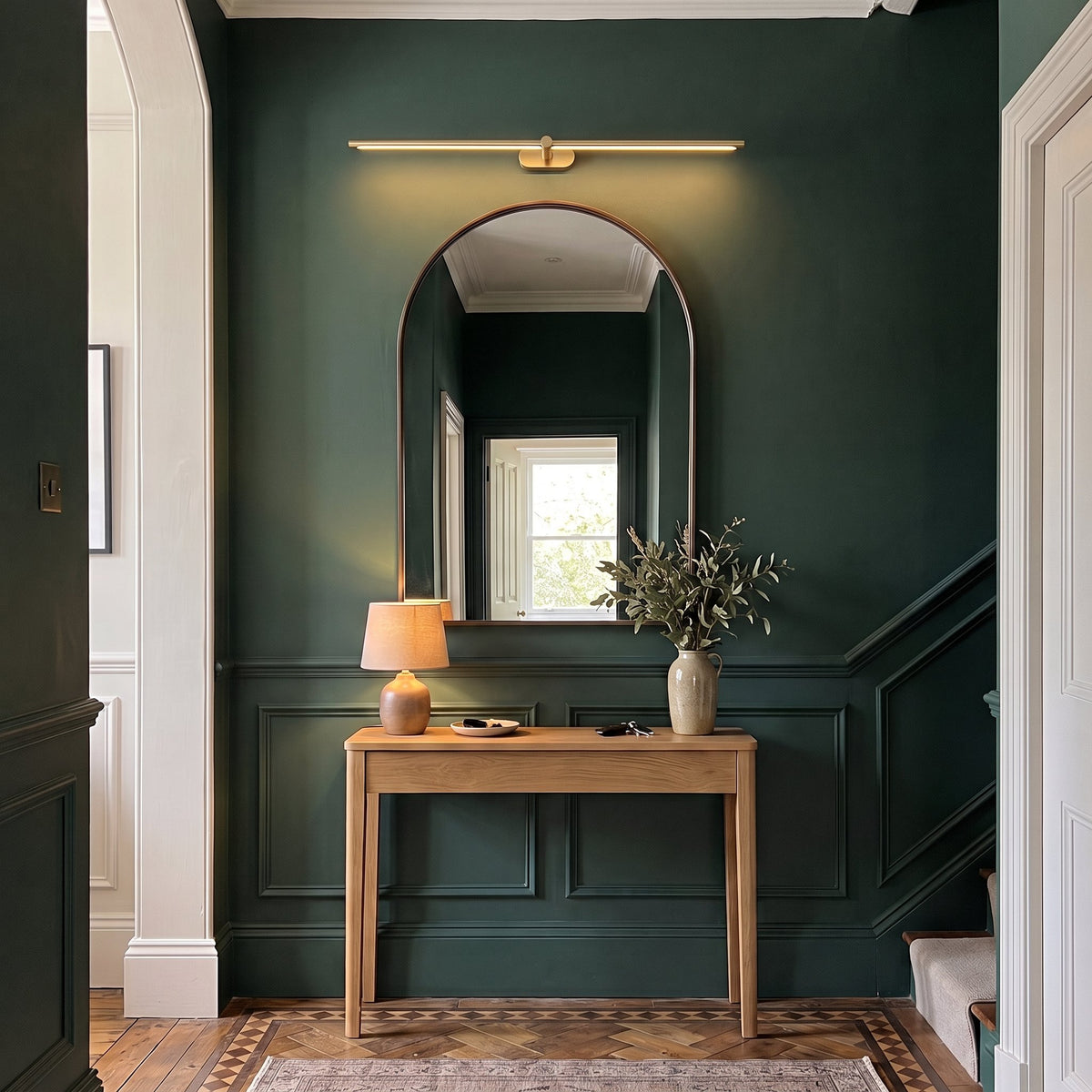 Doven gold-finish linear LED wall light above an arched mirror and oak console in a deep green British hallway