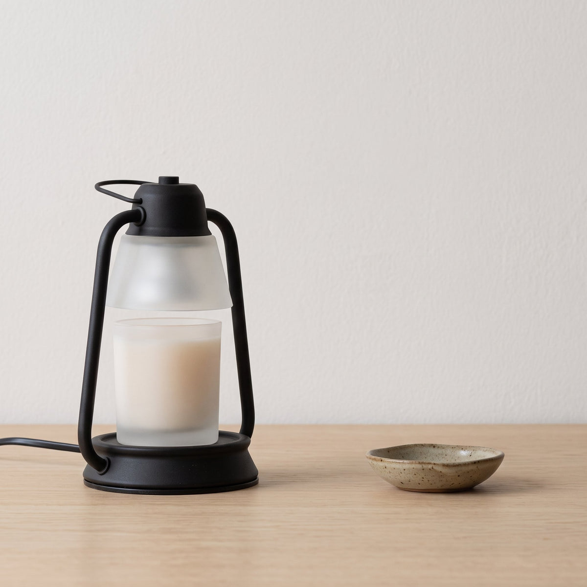 Embra black lantern candle warmer lamp with a frosted candle jar on an oak table beside a ceramic dish