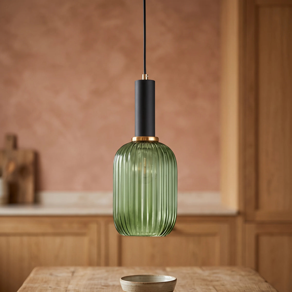 Kael green ribbed glass cylinder pendant light with a black cap against a terracotta clay kitchen wall and oak joinery