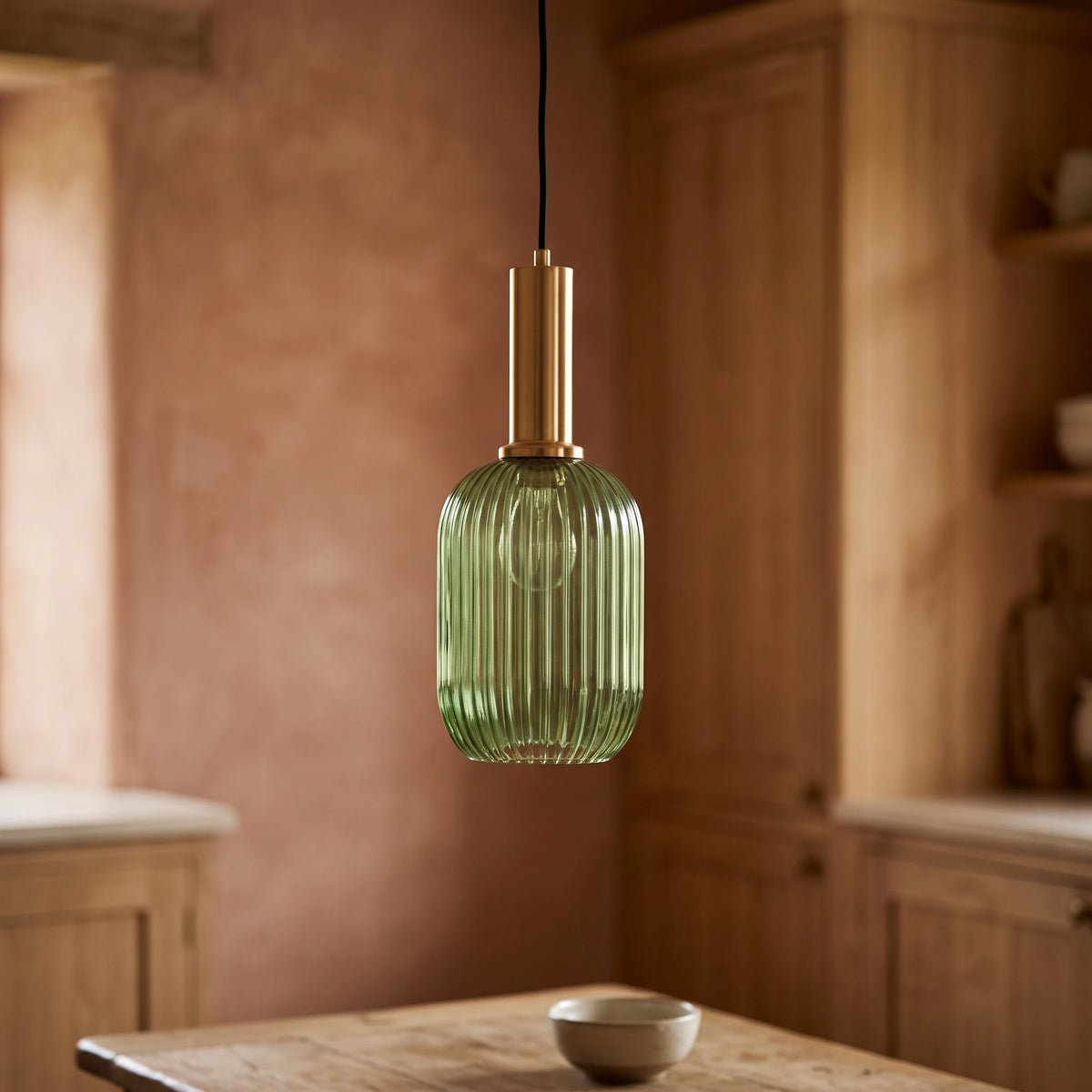 Kael green ribbed glass cylinder pendant light with a brass cap against a terracotta clay kitchen wall and oak joinery