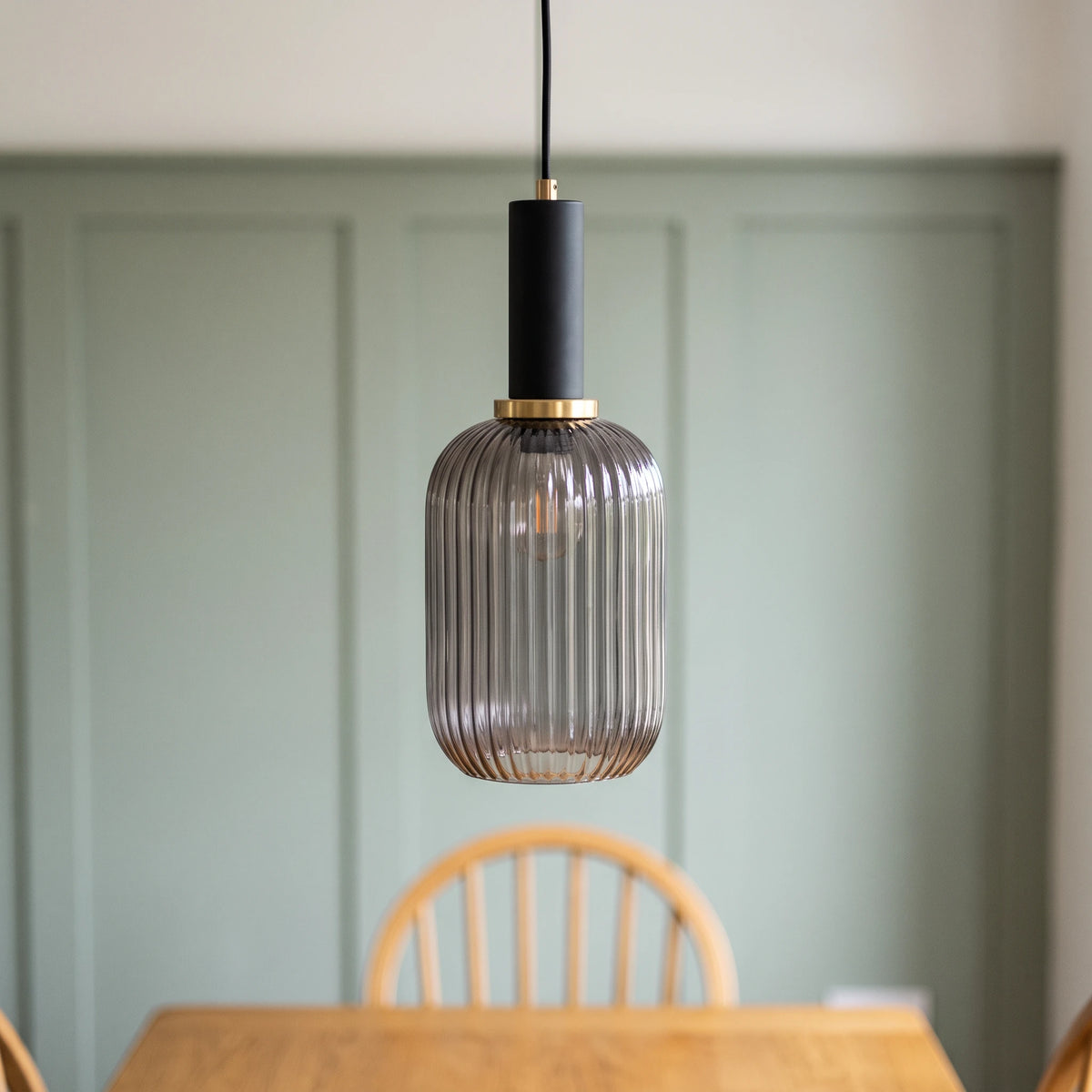 Kael smoked grey ribbed glass cylinder pendant light with a black cap against a sage green panelled dining nook wall