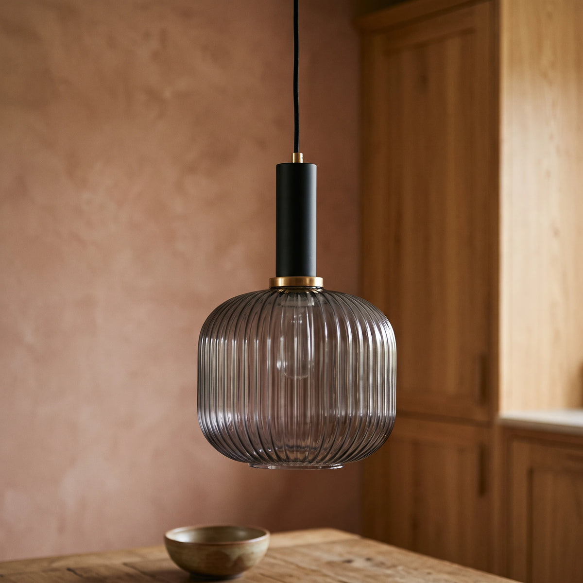 Kael smoked grey ribbed glass jar pendant light with a black cap against a terracotta clay kitchen wall and oak joinery