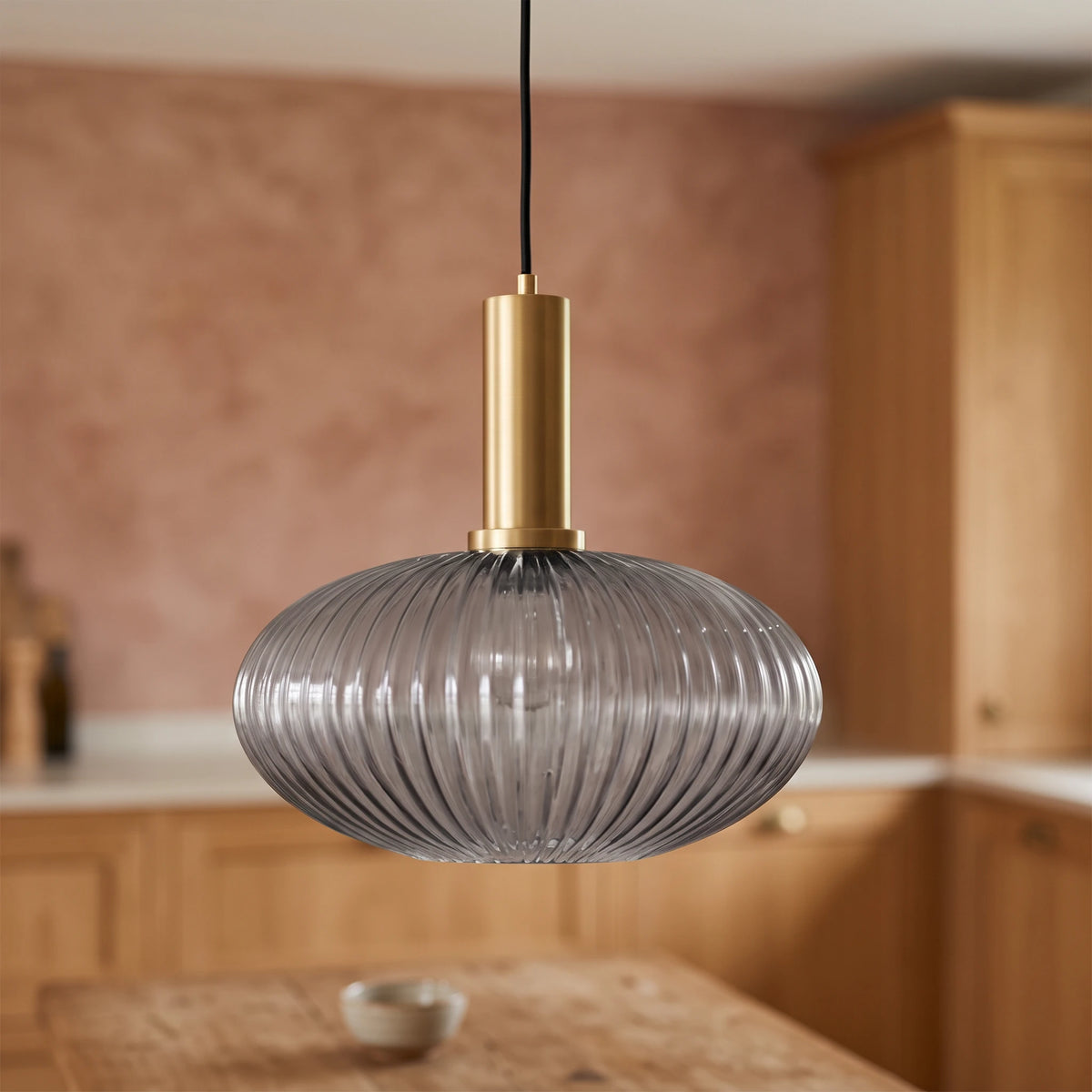 Kael wide smoked grey ribbed glass pendant light with a brass cap against a terracotta clay kitchen wall and oak joinery