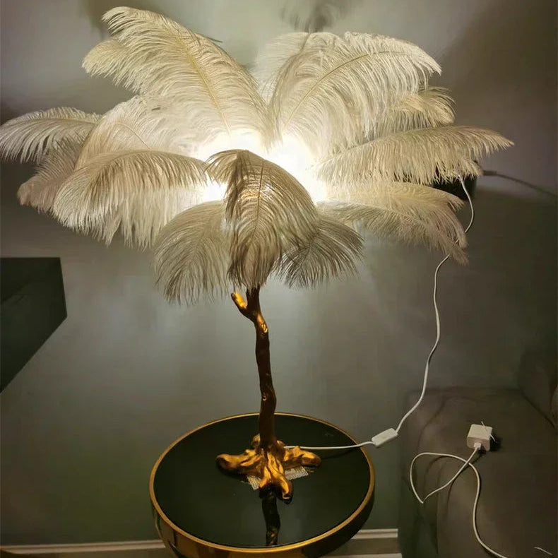 Lorne white ostrich feather lamp with a gold branch base on a black round table with its white cable and plug