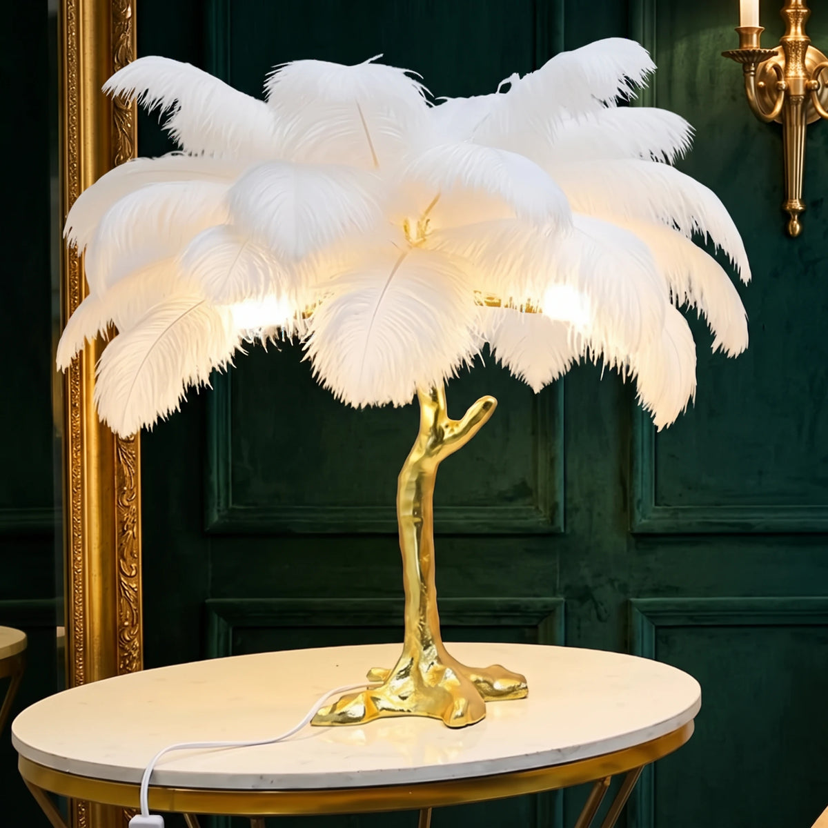 Lorne white ostrich feather lamp with a gold branch base on a marble side table against a deep green velvet wall