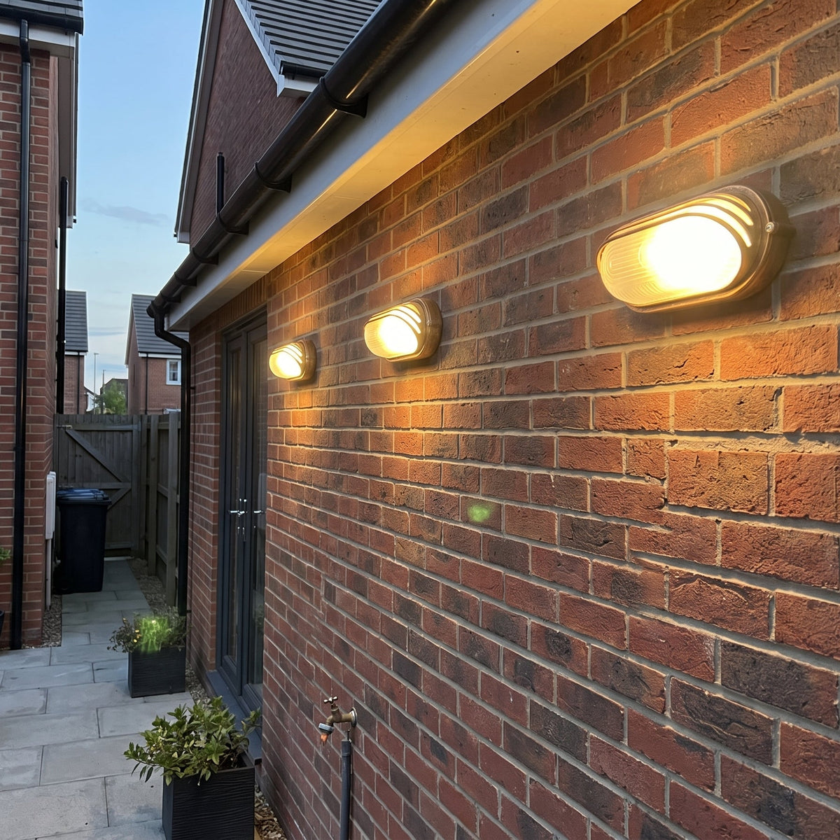 Three Lude bulkhead wall lights lit along a brick wall beside a path