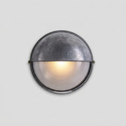 A round industrial-style outdoor wall light with a textured metallic dome and a glowing frosted glass lower half, shown against a light grey background.