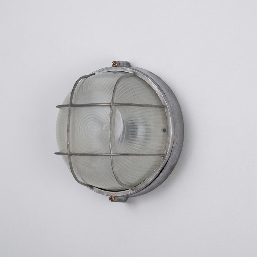 An industrial-style round outdoor wall light fixture with a ribbed clear glass shade and a weathered silver metal frame and cage, mounted on a plain white wall.