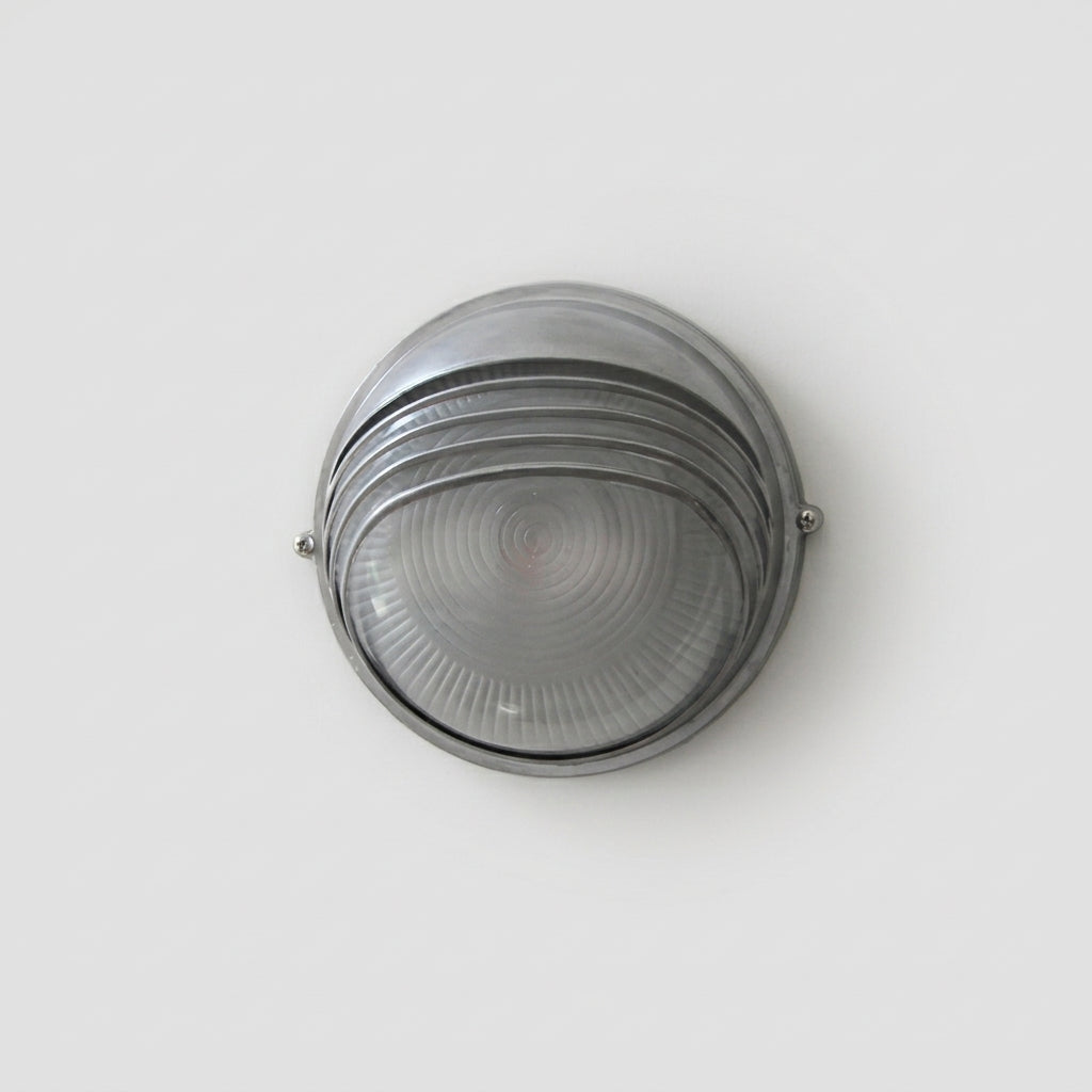 A round industrial wall light fixture with a brushed silver metal frame and a ribbed clear glass shade, shown against a plain light grey background.