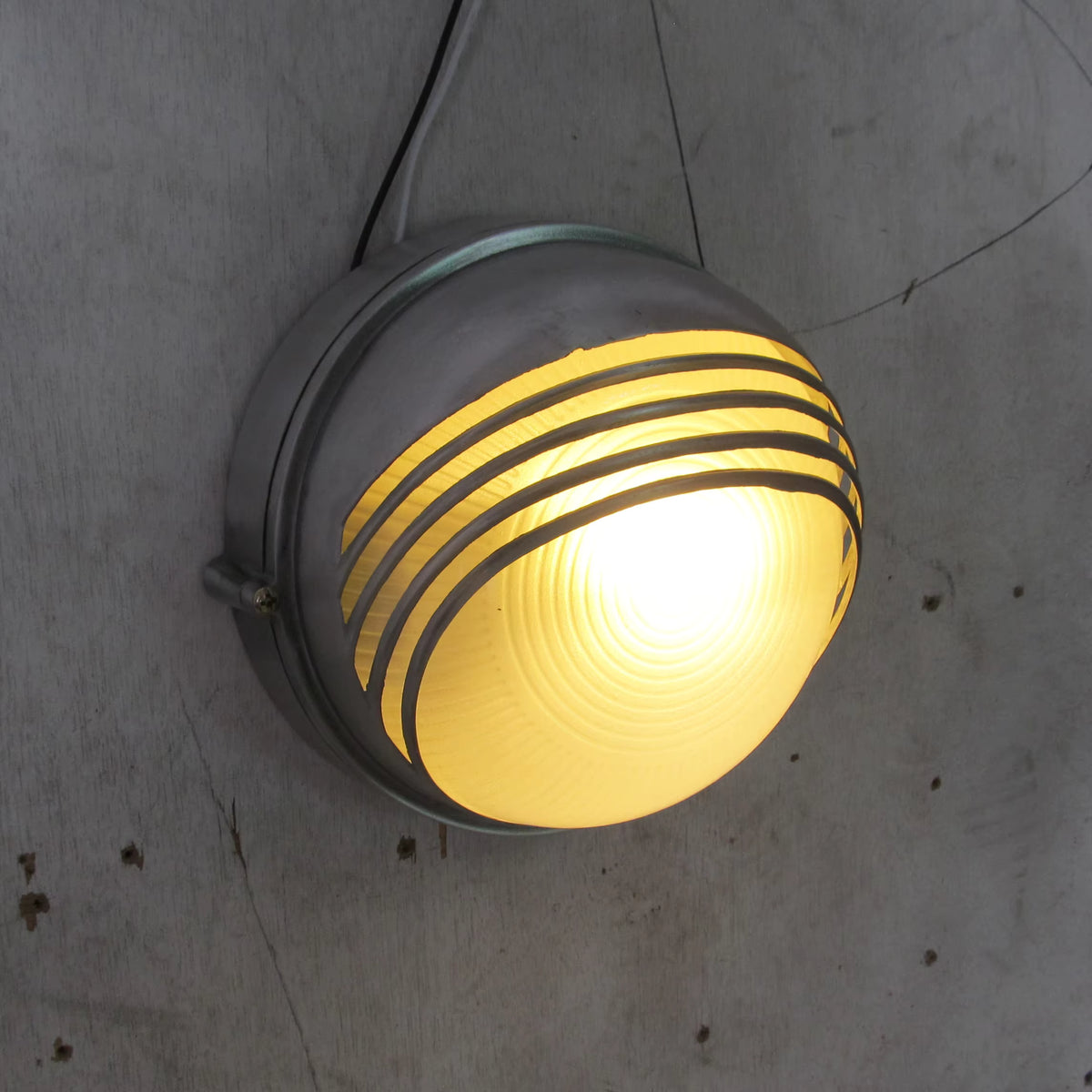 A vintage industrial-style wall light fixture with a ribbed yellow glass globe and a grey metal cage, mounted on a textured concrete wall.