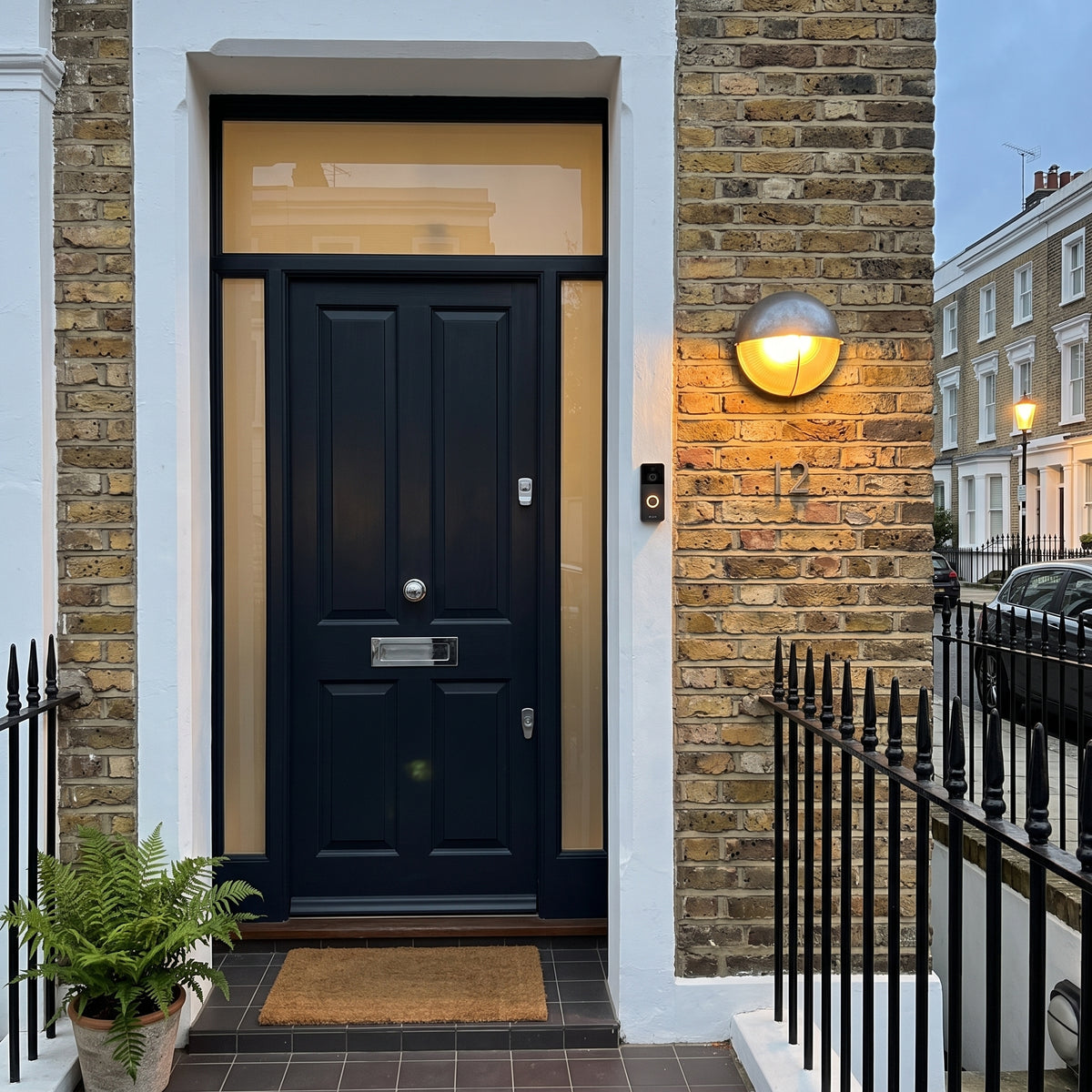 A modern black front door with a glass transom and brass letterbox, set in a white-framed entryway with a brick wall and a spherical wall light. A small potted fern sits at the bottom left.