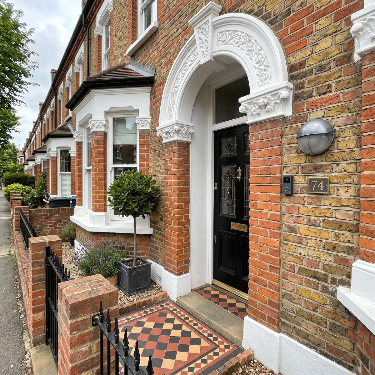 A classic red brick terraced house frontage with a black front door, silver wall light, white decorative archway, tiled entryway, small potted tree, and black metal fence.