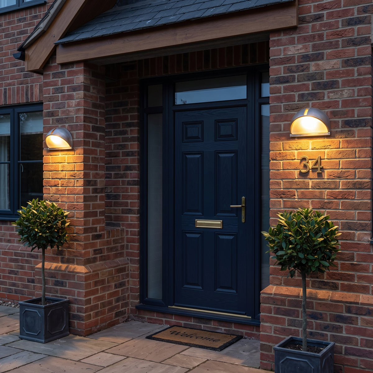 A dark navy blue front door with brass hardware, set in a red brick house with two wall-mounted lights, potted plants, and a 'Welcome' doormat.