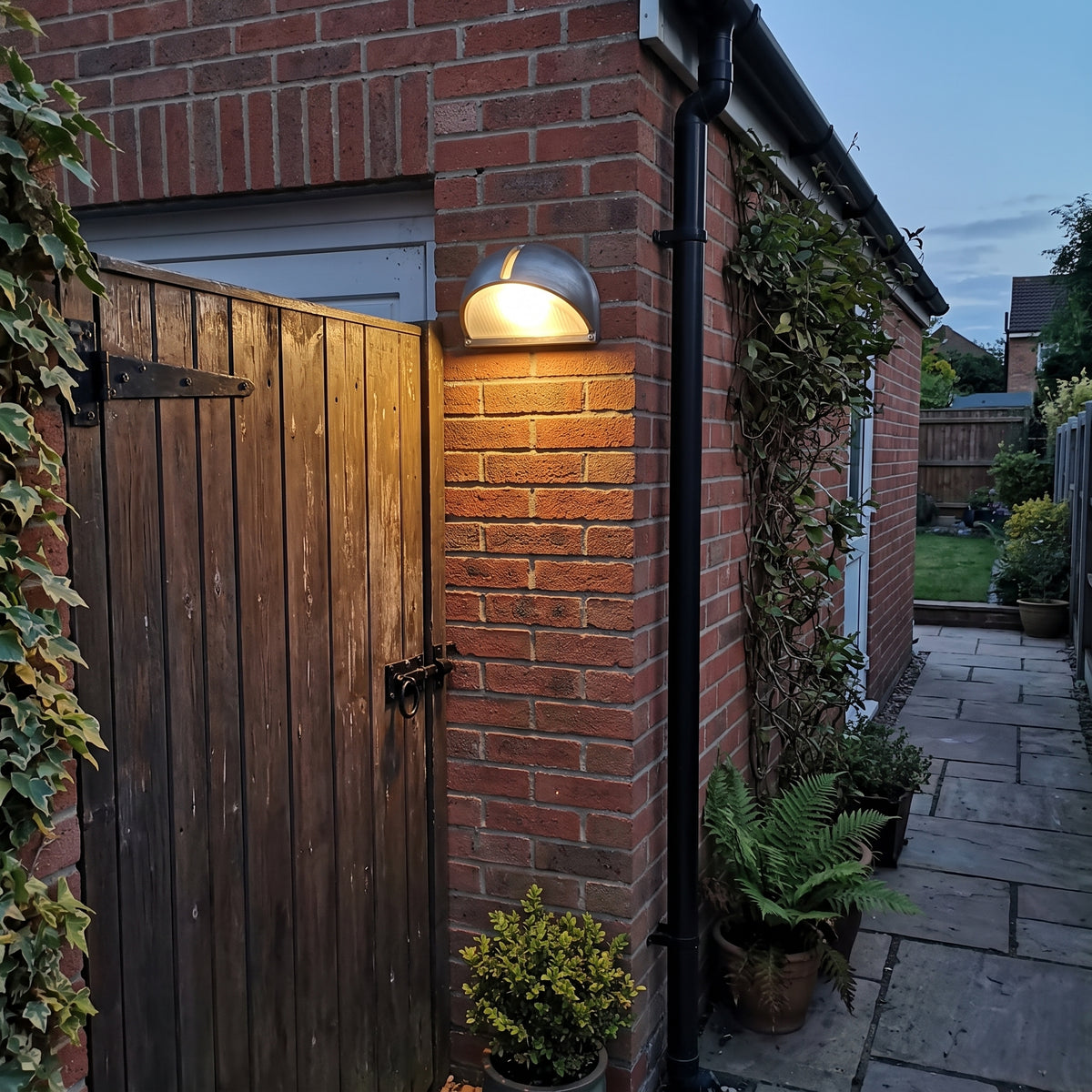 A modern dome-shaped outdoor wall light mounted on a red brick wall next to a wooden gate, casting a warm glow in a backyard setting.
