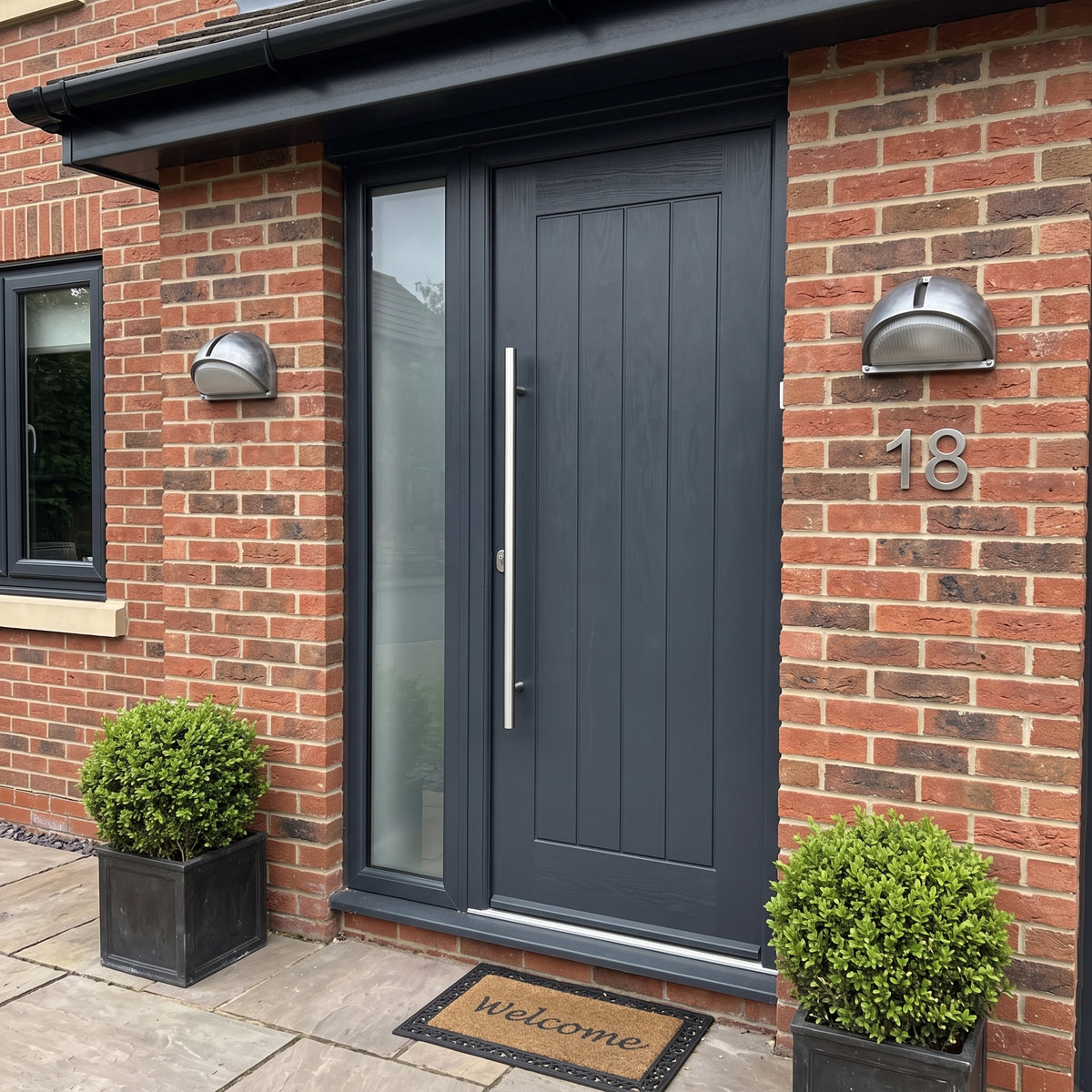 A modern dark gray composite front door with a vertical silver handle, installed in a red brick house. The door has a tall glass panel on the left and a white welcome mat on the threshold with a wall lights fitted on each side of the doors.