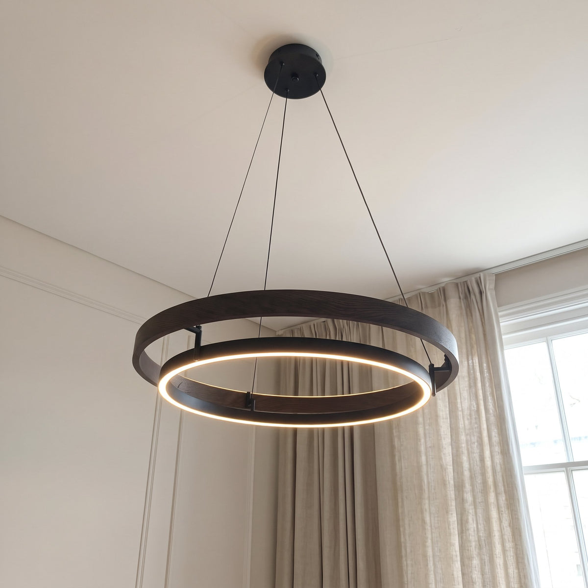 Nyles two-ring wood-grain LED chandelier hanging beside a linen curtain in a neutral room