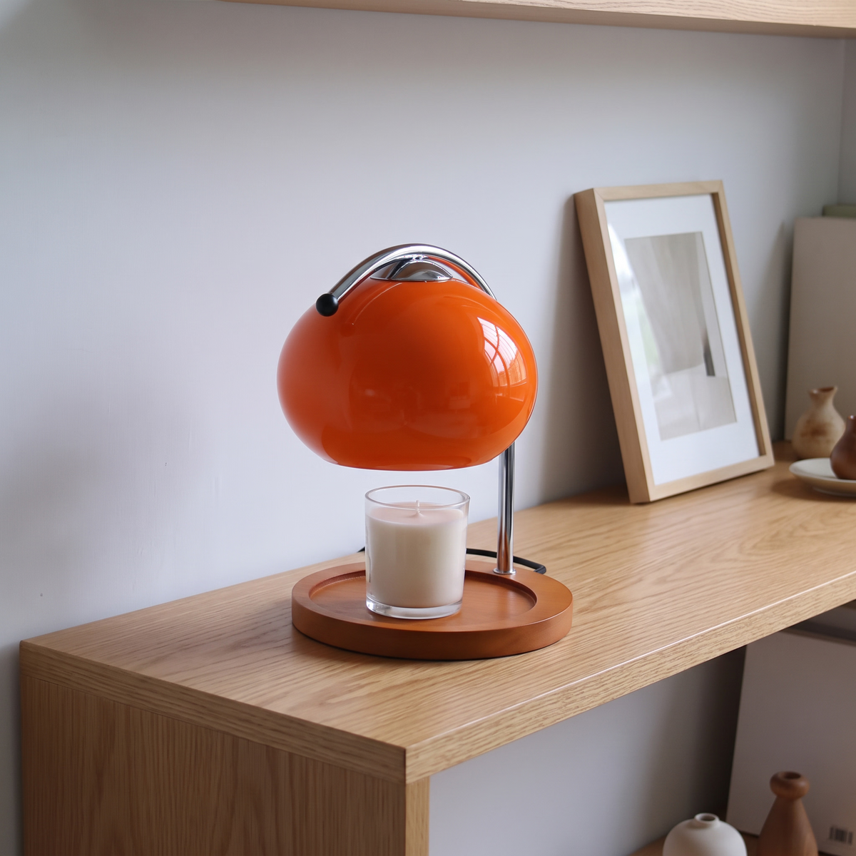 Ondo orange glass candle warmer lamp on a light oak shelf in front of a framed print