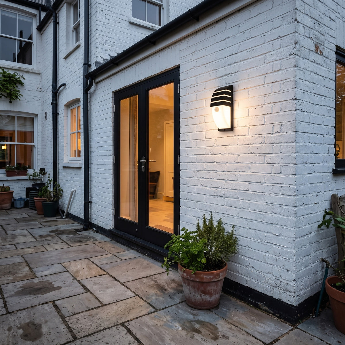 Small Strata outdoor wall light beside black patio doors on a white brick wall in the evening