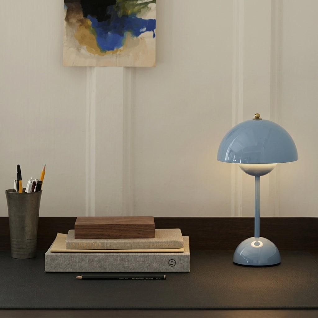 A light blue sulba mushroom-shaped table lamp with a warm glowing bulb, styled on a dark desk next to a pencil cup and books against an off-white paneled wall.