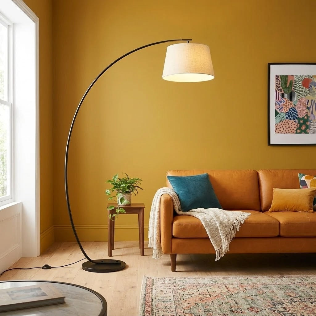 Tavio arc floor lamp with a flaxen fabric drum shade arching over a tan leather sofa in a living room with a mustard wall