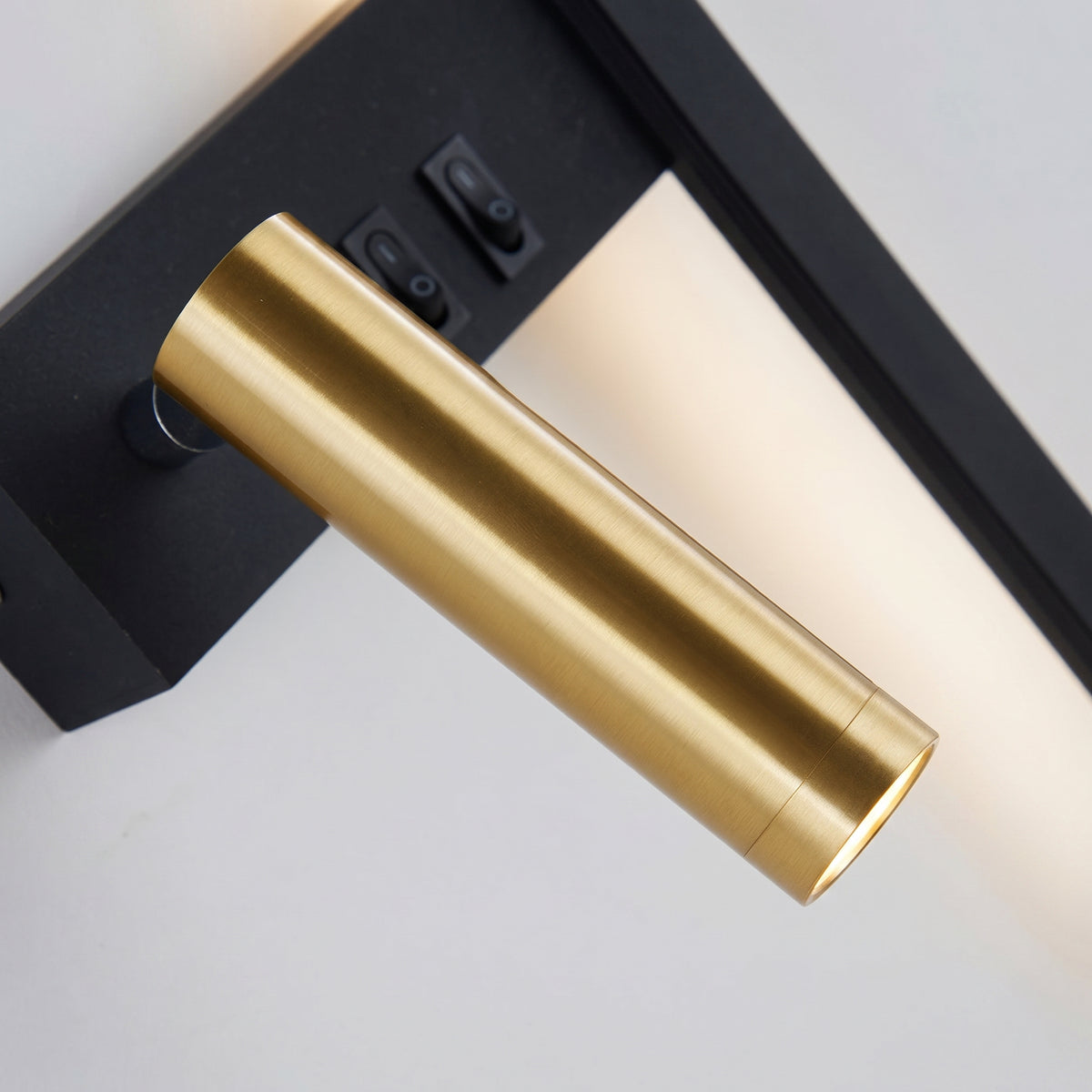 A close-up of a modern veqi bedside wall sconce featuring an adjustable, brushed gold cylindrical LED reading spotlight mounted on a matte black backplate with two built-in toggle switches for independent light control