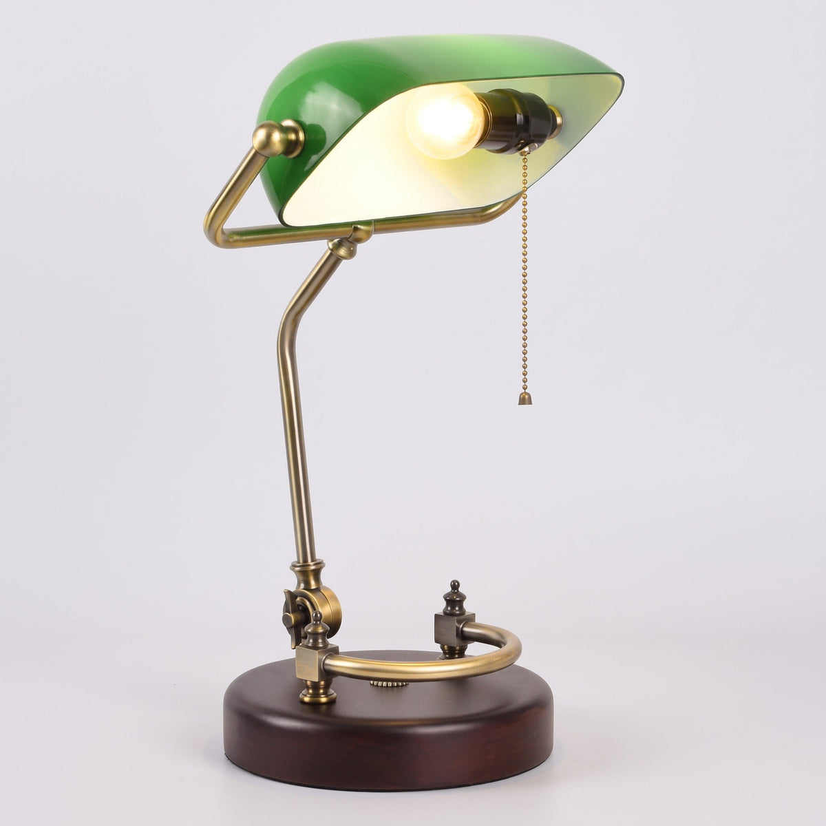 Verda green glass bankers lamp switched on with the bulb visible under the shade, on a light grey studio background